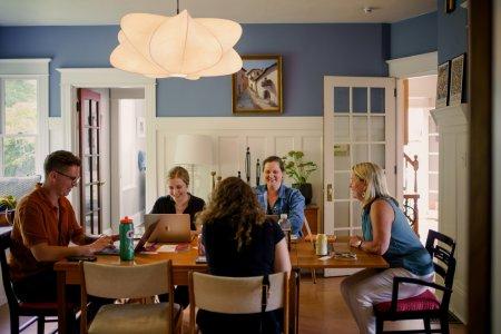 5 adults sitting at a table all focused on their individual laptops. It would seem something amusing or funnay was just said, as everyone has a notable smile on their face.