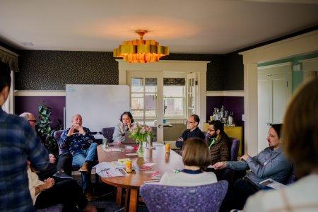 A diverse group of people sitting around a table. All people are focused on a single person who is talking while gesturing with his hands.