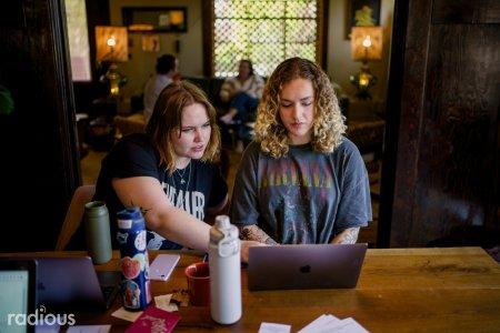 2 adults sitting at a table focused on a single laptop. One of the people is pointing to something on the laptop.